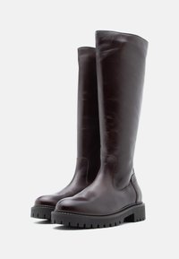 Botas de couro castanho-escuro até aos joelhos, com bicos arredondados e solas grossas em preto, apresentadas sobre um fundo claro e simples.