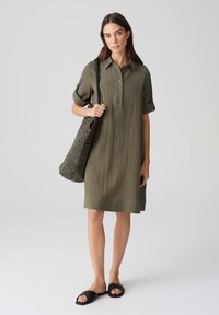 Abito verde oliva con colletto, maniche corte arrotolate, texture a coste e chiusura con bottoni. Abbinato a una borsa tote nera intrecciata e sandali.