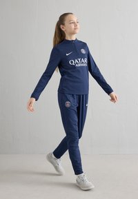 Tengerészkék sportos szett félcipzáros felsővel és hozzáillő nadrággal, melyeken fehér logók: Nike és Qatar Airways láthatók. Világosszürke tornacipővel viselve.