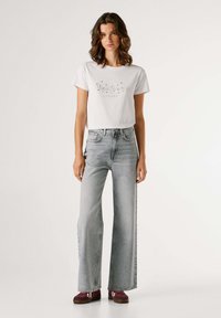 Camiseta blanca de manga corta con un diseño estampado, combinada con vaqueros de tiro alto, pierna ancha y color gris claro, y zapatillas deportivas color granate.