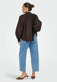Camicia oversize marrone con maniche a palloncino, colletto alto e polsini con bottoni; abbinata a jeans a gamba larga blu chiaro e décolleté marroni.