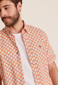 Korte mouwen shirt in een oranje, gele en blauwe bloemenpatroon; met knopen aan de voorkant; gedragen over een wit T-shirt; de stof lijkt lichtgewicht.