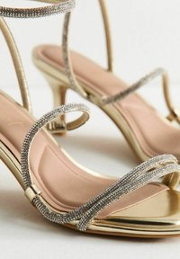 Goldene High Heels Sandalen mit dünnen Riemen, die mit kleinen Strasssteinen verziert sind, und einem hautfarbenen gepolsterten Fußbett auf weißem Hintergrund.