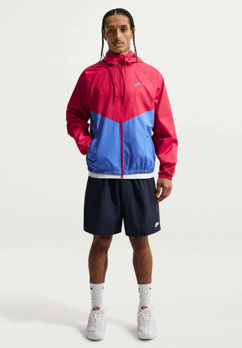 Man met gevlochten haar, een rood-blauw Nike jack met rits, zwarte Nike shorts, witte sokken en witte sneakers, staand met handen in de zakken.