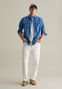 Blauw gestreepte overhemd met knopen, een wit T-shirt eronder, witte broek en bruine mocassinschoenen, met een blauw-wit gestreepte pet.
