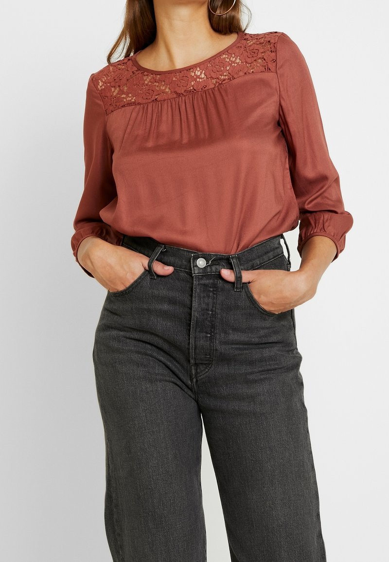 Blusa color ruggine con dettaglio in pizzo sul colletto, maniche lunghe e vestibilità rilassata, abbinata a jeans in denim nero a vita alta.