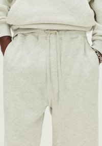 Light gray sweatpants with drawstring waist, side pockets, and hands partially in pockets, paired with matching sweatshirt sleeve and silver bracelet.