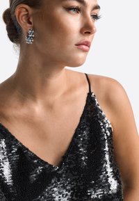 Čierna satičková top s tenkými ramienkami. Disponuje hlbokým výstrihom do V a lesklou textúrou. Strieborné kruhové náušnice dotvárajú vzhľad.