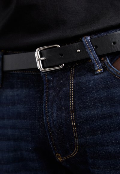 Ceinture en cuir noir avec une boucle rectangulaire en argent, portée sur un jean en denim foncé avec des coutures jaunes. Texture lisse, détails minimaux.