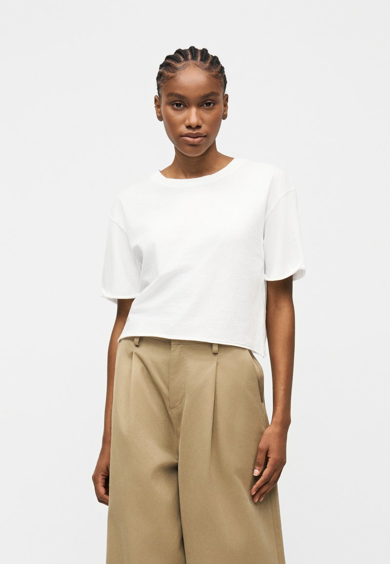 Νεαρή γυναίκα με πλεξούδες, που φοράει απλό λευκό cropped μπλουζάκι με κοντά μανίκια και μπεζ ψηλόμεσα παντελόνια με φαρδιά πόδια.