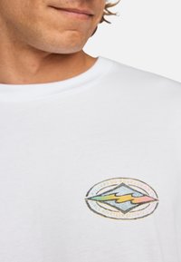 Camiseta de algodón blanca con un logo gráfico redondo y multicolor que presenta montañas y olas, bordeado por texto en negro y amarillo.