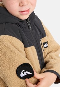 Chaqueta con cremallera en marrón y negro, hecha de material de forro polar, con un cuerpo texturizado en tono beige, mangas negras lisas y un parche de logo prominente en la manga.