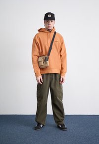 Orange hættetrøje med kængurulomme, sort kasket, olivengrøn cargo-bukser, og en lille tan crossover-taske. Sorte løbesko med hvide striber.