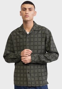 Cămașă verde cu un model, având guler, nasturi în față, mâneci lungi și un design în rețea de culoare neagră. Material moale, texturat.