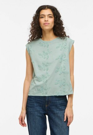 Donna con capelli ricci e scuri che indossa un top senza maniche verde salvia con ricami floreali e jeans blu, in piedi davanti a uno sfondo bianco.