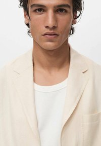 Lys beige blazer over en hvid ribbet skjorte. Blazeren har et minimalistisk design, klapkraver og en brystlomme med en blød tekstur.