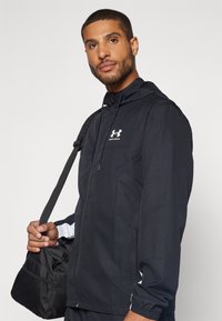 Čierna vetrovka s predným zipsom, nastaviteľnou kapucňou a elastickými manžetami; obsahuje biele akcenty a logo na hrudi.