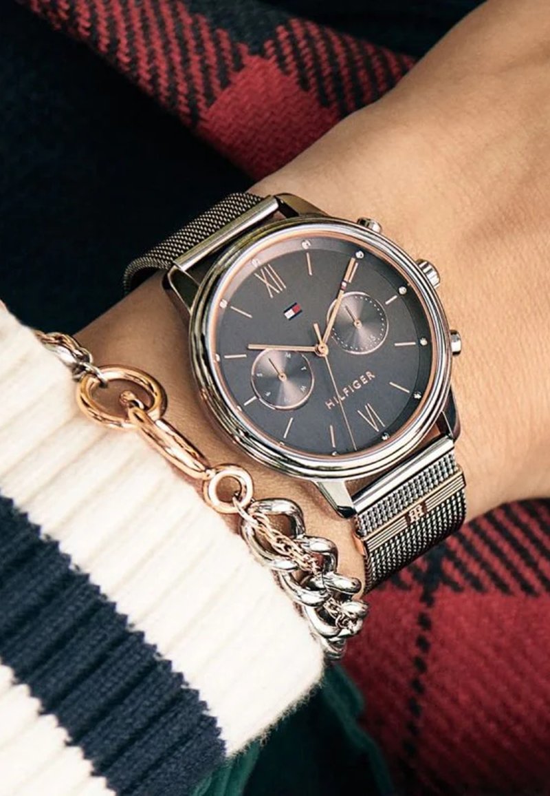 Montre avec bracelet en maille argentée, cadran gris foncé, chiffres romains et deux sous-cadrans. Mise en valeur par un accent en or rose au niveau de la lunette et du fermoir.