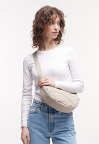 Beige qviltet taske med en bred rem, let materiale, dråbeformet og diskret branding, båret crossbody af en model.