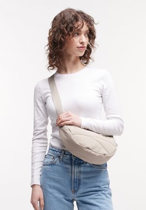 Bolso de mano acolchado beige con una correa ancha, material ligero, forma de gota y branding sutil, llevado como bandolera por un modelo.