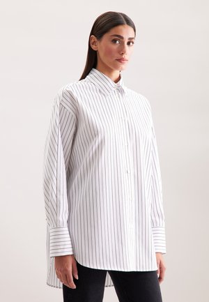 Donna che indossa una camicia bianca a maniche lunghe oversize con strisce verticali nere, abbottonata, abbinata a pantaloni neri, su uno sfondo semplice.