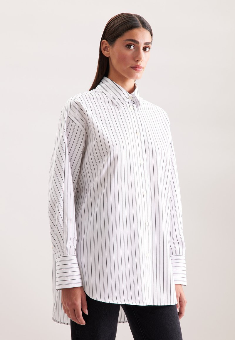 Vrouw in een wit oversized overhemd met lange mouwen met zwarte verticale strepen, dichtgeknoopt, gecombineerd met zwarte broek, tegen een effen achtergrond.