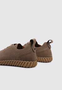 Des chaussures en daim marron avec une surface texturée, un design perforé, un système de laçage à plat et une semelle en caoutchouc flexible avec des rainures horizontales.