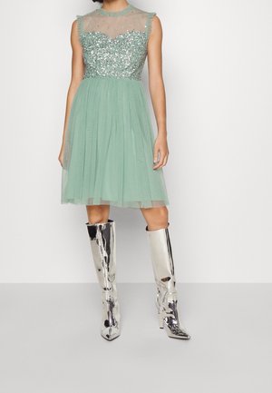 Femme portant une robe verte sans manches avec un corsage à paillettes et une jupe en tulle, associée à des bottes argentées brillantes à talons montant jusqu'aux genoux sur un sol gris.