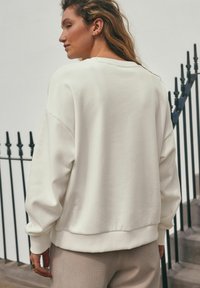 Biała bluza z luźnym krojem, ściągaczami na rękawach i okrągłym dekoltem. Miękka tekstura i minimalistyczny design, noszona na fakturowanych beżowych spodniach.