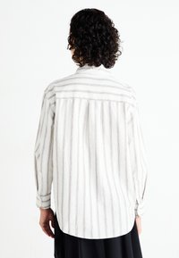 Chemise boutonnée blanche avec des rayures brun clair, dotée d'un empiècement au dos, de plis et de poignets. Le tissu semble léger et texturé.