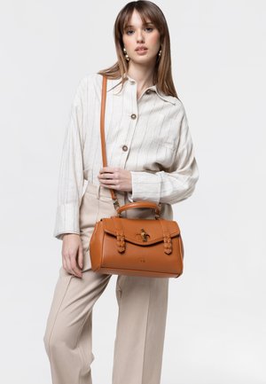 Giovane donna con capelli lunghi che indossa pantaloni beige e una camicia a righe, che tiene in mano una borsa in pelle marrone chiaro con tracolla.