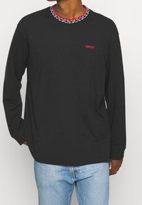 Person, der ein schwarzes Levi's Langarmhemd mit einem rot-weißen gemusterten Kragen trägt, kombiniert mit hellblauen Jeans, vor einem einfarbigen Hintergrund.