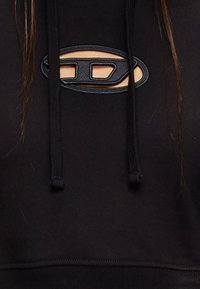Zwarte hoodie met een getextureerd logo in een uitsnijdvorm, met trekkoorden en een zachte stoftextuur. Geen aanvullende details zichtbaar.
