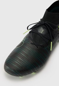 Black football boot with a sleek design, featuring green accents and textured lines across the upper. Includes black laces and a sock-like collar.