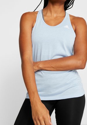 Kobieta w jasnoniebieskim bezrękawniku sportowym Adidas i czarnych legginsach, z jedną ręką skrzyżowaną na talii.