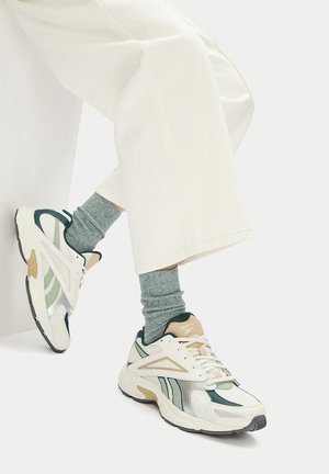 Reebok sportiniai bateliai su balta tinkliniu viršumi, žaliais ir sidabriniais akcentais, kreminiu padu ir pilkomis tekstūruotomis kojinėmis.