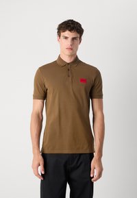 Polo din bumbac maro, cu mâneci scurte, plachetă cu trei butoane, guler ribat și un patch cu logo roșu pe partea stângă a pieptului.