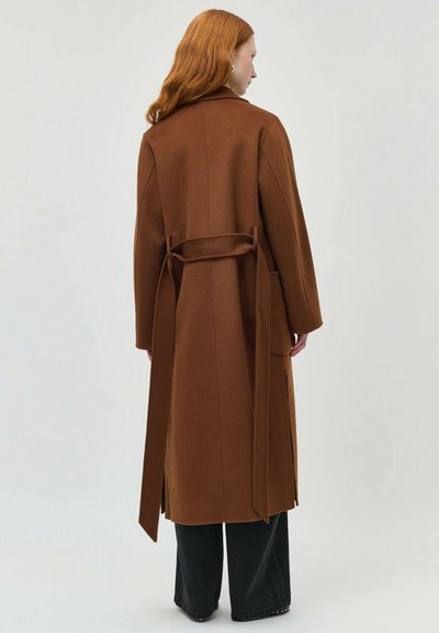 Femeie cu păr roșu, purtând o haină lungă maro cu curea și pantaloni negri largi, stând cu fața spre spate, pe un fundal simplu.