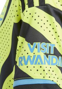 Jersey deportivo de color amarillo y negro con un patrón que presenta "VISIT RWANDA" en azul. El diseño incluye puntos y un ribete azul contrastante en la parte inferior.