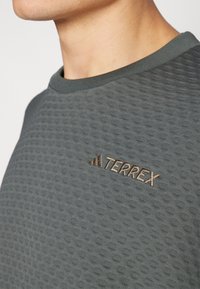 Chemise grise texturée avec un col rond. Présente un motif en relief et un logo "TERREX" brodé marron sur la poitrine.