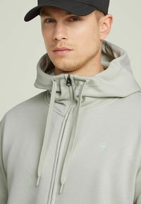 Haut de sweat à capuche zippé gris clair avec un col montant, cordons de serrage ajustables et un petit logo. Tissu lisse avec une coupe décontractée. Casquette noire portée au-dessus.