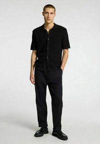 Camicia nera a maniche corte con bottoni in tessuto strutturato, abbinata a pantaloni neri larghi e scarpe nere stringate. Design semplice e moderno.