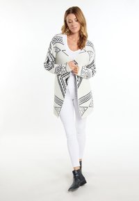 Strickjacke in Creme mit schwarzen geometrischen Mustern, offenes Design, lange Ärmel und mittellange Form, getragen über einem weißen Top und weißen Hosen.