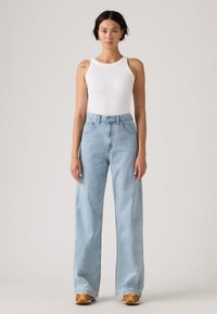 Νεαρή γυναίκα, βλέποντας μπροστά, φοράει λευκή μπλούζα χωρίς μανίκια, ανοιχτόχρωμα μπλε τζιν με φαρδιές παντελόνες και κίτρινα παπούτσια.