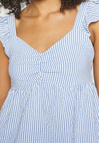Femme portant une robe à rayures verticales bleues et blanches avec un col en V froncé et des manches courtes à volants.