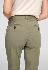Persona che indossa pantaloni verde oliva con tasche posteriori abbottonate e una camicia beige infilata, mostrata dalla vita in giù.