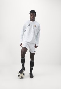 Hvid langærmet fodboldtrøje med et geometrisk mønster, hvide shorts, sorte strømper med hvide striber og sorte fodboldstøvler, stående på en fodbold.