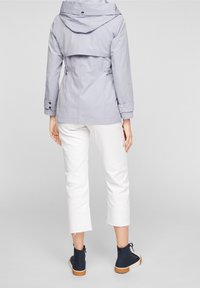 Veste imperméable gris clair avec capuche, boutons-pression et taille ajustable. Associée à un pantalon blanc court et des chaussures montantes bleu foncé.