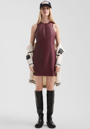 Kobieta w bezrękawowej sukience ze skóry w kolorze bordo, czarnych kozakach sięgających kolan, czapce w kolorze czarnym oraz beżowym kardiganie zarzuconym na ramiona na białym tle.