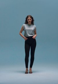 Grijze mouwloze top gecombineerd met hoge zwarte skinny jeans en zwarte open teen hakken, tegen een zachte blauwe achtergrond.